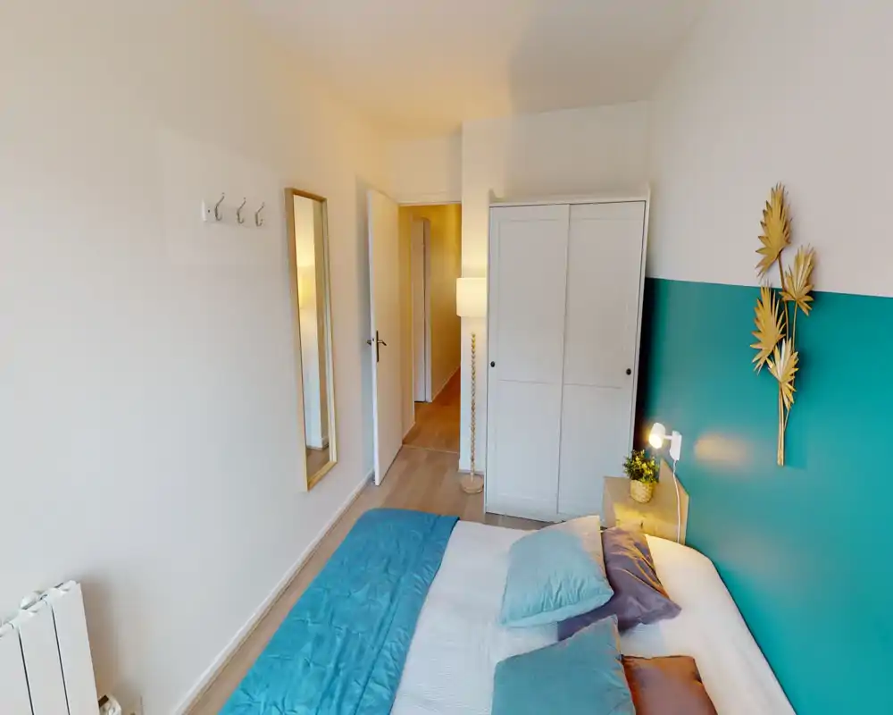 Bright, tidy bedroom with a turquoise accent wall and a double bed dressed in coordinated pillows and a quilt. A white wardrobe, full-length mirror and bedside lamp complete the calm, welcoming layout.