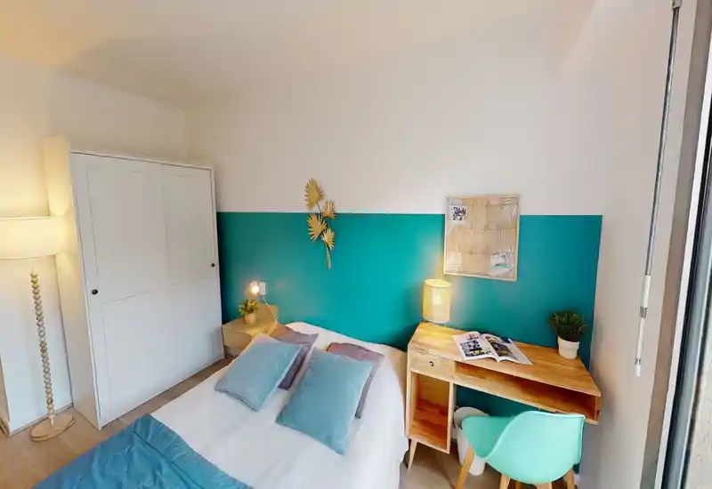Bright, cozy bedroom with a teal accent wall, double bed dressed with cushions and a wooden desk by the window — well-lit and stylish for a comfortable stay.