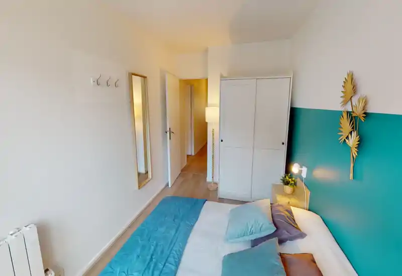 Bright, tidy bedroom with a turquoise accent wall and a double bed dressed in coordinated pillows and a quilt. A white wardrobe, full-length mirror and bedside lamp complete the calm, welcoming layout.