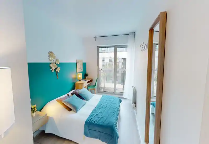 Bright, modern bedroom with a double bed dressed in white linen and teal accents, a bedside lamp and small wooden desk beside large floor-to-ceiling windows letting in generous natural light.
