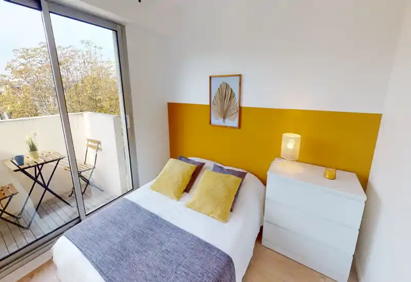 Bright, stylish bedroom with a double bed, decorative cushions and a large sliding door leading to a small balcony. The yellow accent wall and bedside lamp create a warm, inviting atmosphere ideal for rentals.