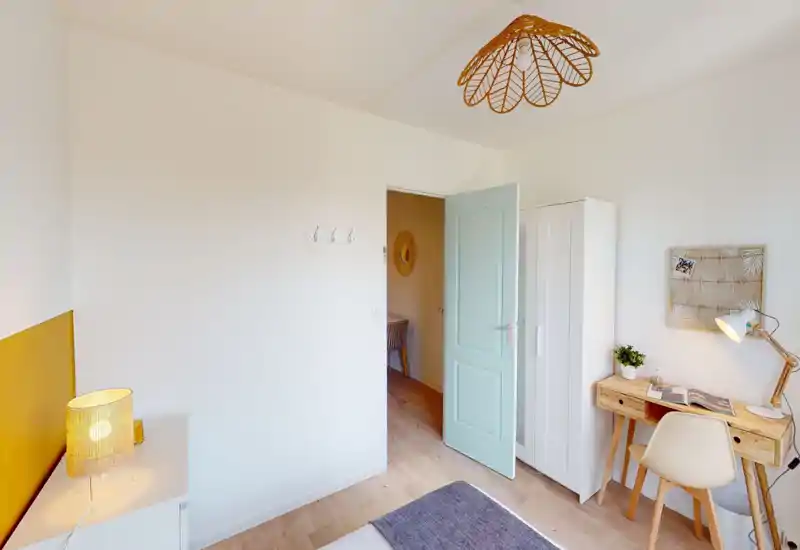 Bright, modern bedroom corner with a visible bed corner, a wooden desk and wardrobe; warm lamp and soft decor create a cozy, airy atmosphere ideal for short-term rentals.