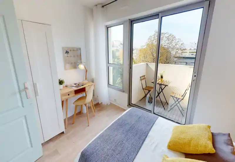 Bright, cozy bedroom with a comfortable double bed, decorative yellow cushions and a small wooden desk next to large sliding glass doors leading to a sunny balcony.