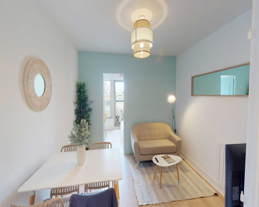 Bright, well-styled living room with a small sofa, coffee table and dining table visible; soft teal accent wall and decorative plants create a calm, welcoming atmosphere.