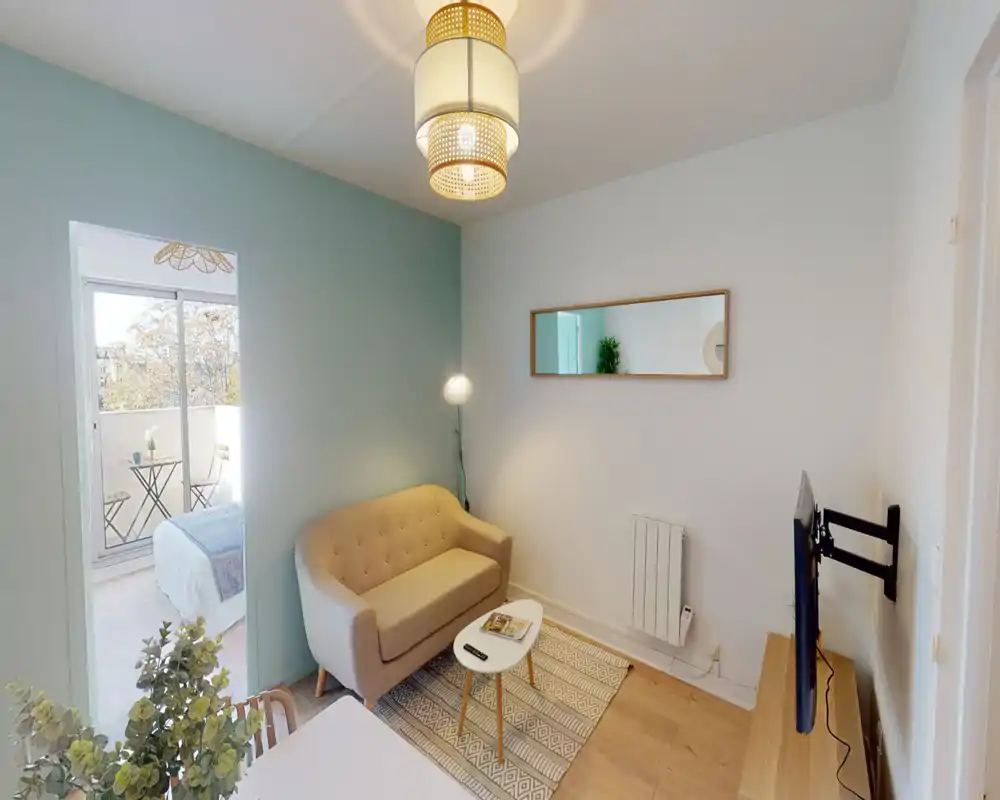 Bright, compact living room with a small beige sofa, patterned rug and a wall-mounted TV — soft pastel accent wall and natural light from the balcony make the space feel airy and inviting.
