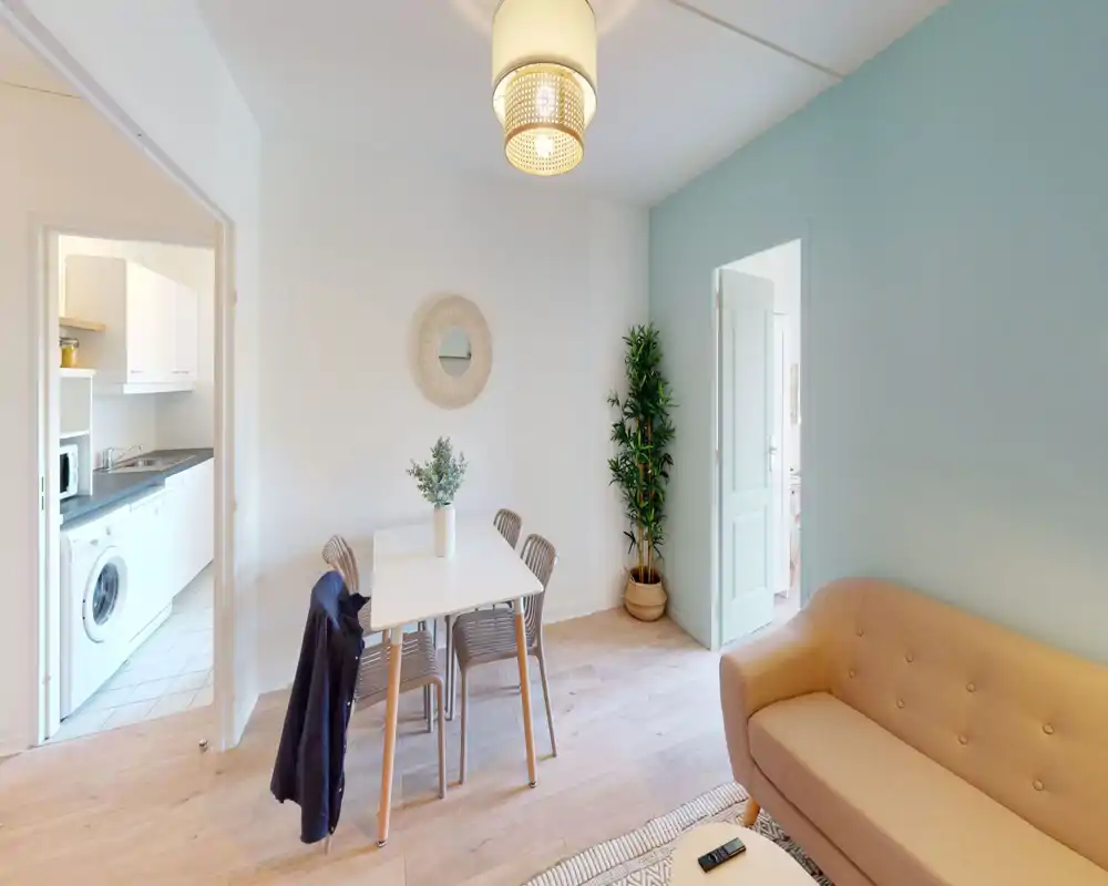 Bright, styled living area with a soft two-seater sofa, a small dining table and chairs, decorative plant and pleasant teal accent wall — a cozy space ideal for relaxing and dining.