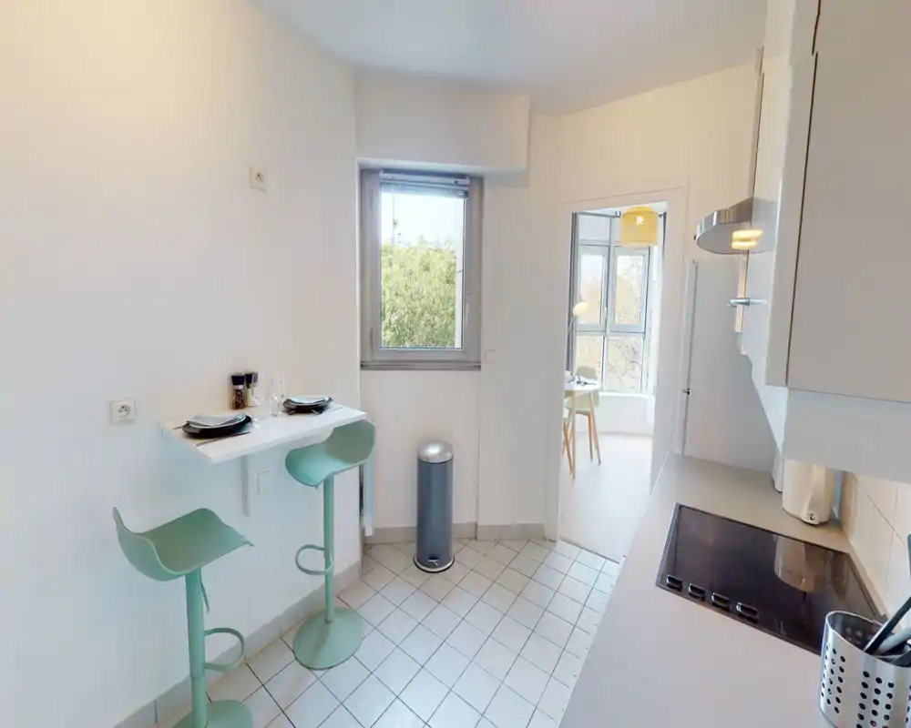 Bright compact kitchen with a built-in electric cooktop, countertop workspace and a small breakfast bar with two mint-green stools — modern, clean and functional for a small apartment.