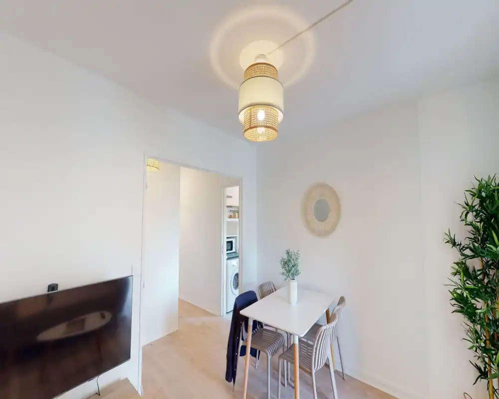 Bright, modern living/dining area with a compact table for four, decorative mirror and a potted plant; warm pendant lighting and a wall-mounted TV create a cozy multifunctional space.