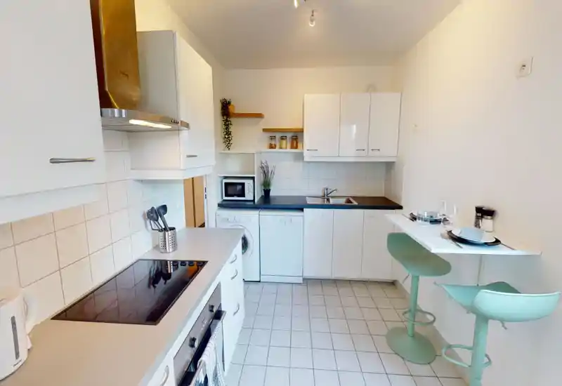 Cuisine moderne et lumineuse avec placards blancs, plaque à induction, évier et bar petit-déjeuner avec tabourets vert menthe ; micro-ondes, lave-vaisselle et machine à laver présents pour un agencement pratique.