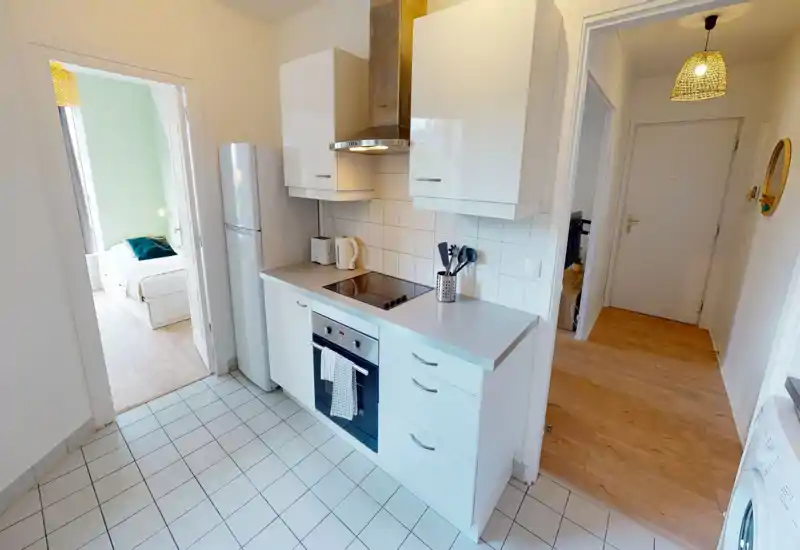 Cuisine moderne et lumineuse avec meubles blancs, plaque à induction, four encastré et réfrigérateur. Sol carrelé propre et plan de travail pratique pour un appartement compact.