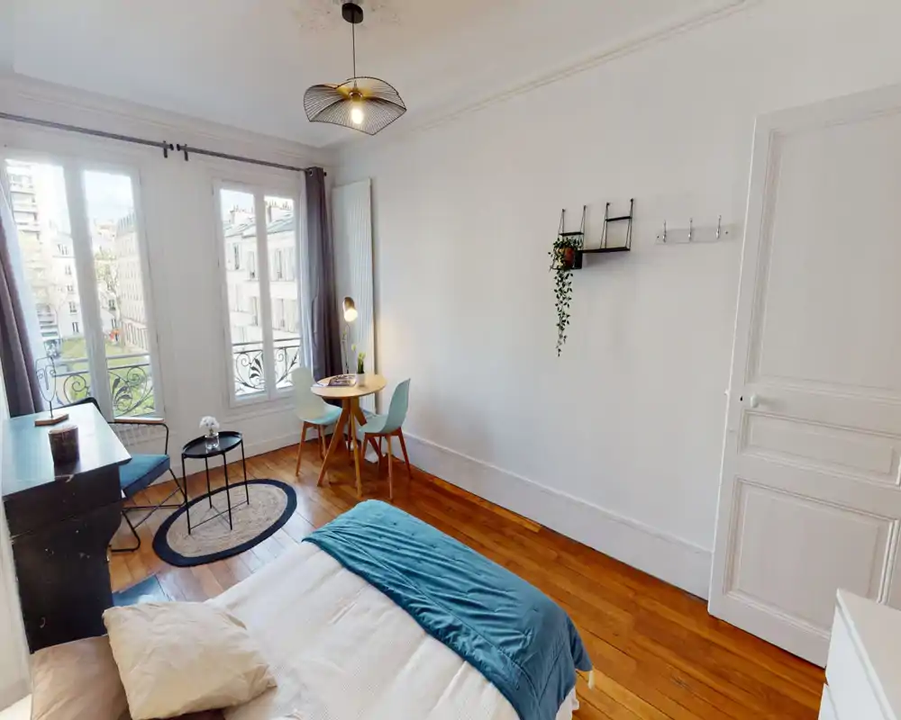 Camera da letto luminosa e curata, con letto matrimoniale, pavimento in legno e grandi finestre che rendono l'ambiente arioso. Tavolino da pranzo e scrivania rendono lo spazio versatile e adatto a soggiorni brevi.
