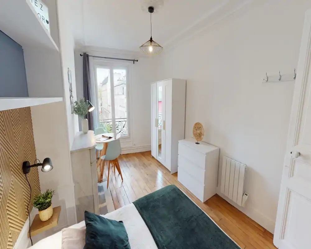 Dormitorio luminoso y bien decorado con la cama en primer plano, suelos de madera, un pequeño escritorio junto a una gran ventana y muebles blancos de almacenamiento — ideal para una persona o pareja.