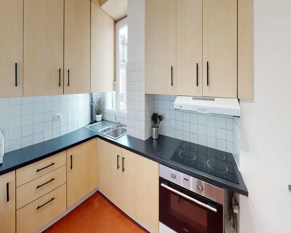 Cucina luminosa e moderna con mobili in legno chiaro, piano di lavoro nero, piano cottura in vetroceramica e forno incorporato; finestra sopra il lavello garantisce luce naturale.