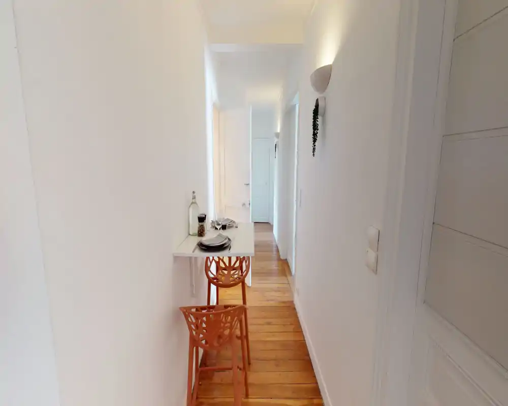 Ingresso/corridoio luminoso e stretto con un piccolo tavolo a parete pieghevole e due sgabelli decorativi, pareti bianche e pavimento in legno caldo che creano un passaggio accogliente.