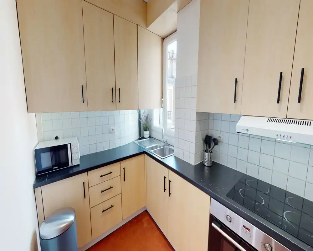 Cucina moderna compatta con mobili in legno chiaro, piani neri, piano cottura elettrico e lavello vicino a una finestra luminosa — ordinata e funzionale per un appartamento.