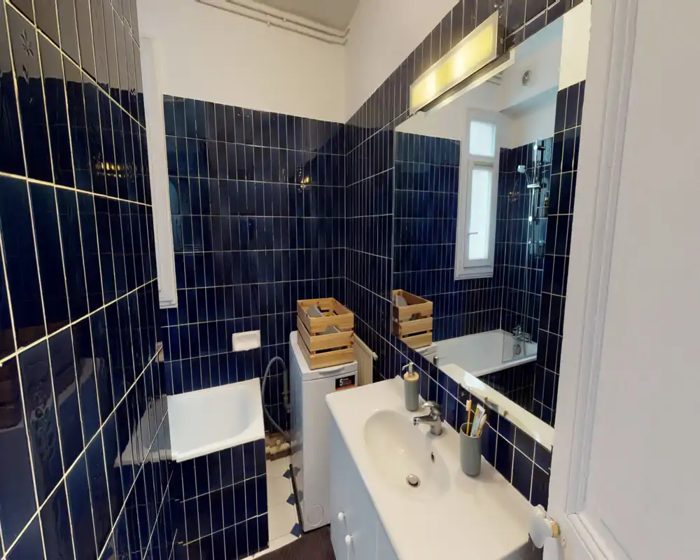Bagno compatto con piastrelle blu scuro, vasca con doccia, lavabo con specchio e una piccola lavatrice; ambiente luminoso e moderno con piastrelle riflettenti.