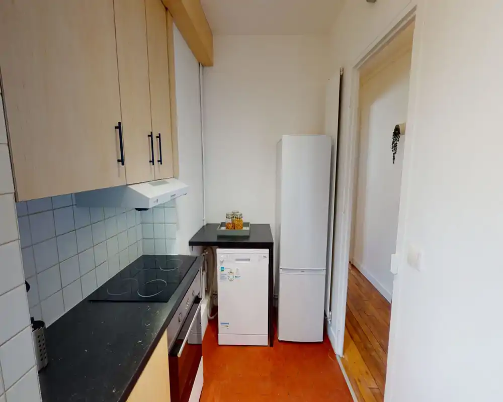 Cucina compatta e luminosa con piano cottura elettrico, forno incassato, lavastoviglie e frigorifero. Pensili in legno chiaro e paraschizzi piastrellato per un ambiente funzionale e pulito.