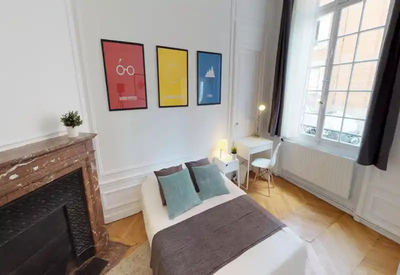 Camera da letto luminosa e di stile con letto matrimoniale al centro, cuscini decorativi e stampe incorniciate sopra. Grande finestra che illumina la stanza e una piccola scrivania per lavorare.