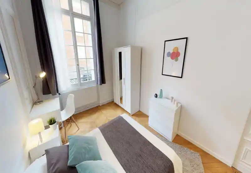 Camera da letto luminosa e elegante con grande finestra, pavimento a spina di pesce in legno e letto matrimoniale ben rifatto con cuscini decorativi e morbido tappeto.