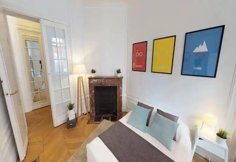 Camera da letto luminosa e di stile con un ampio letto con biancheria neutra, cuscini decorativi e un runner. La stanza ha un camino in marmo classico, quadri alle pareti, parquet caldo e lampade da comodino che creano un'atmosfera accogliente.