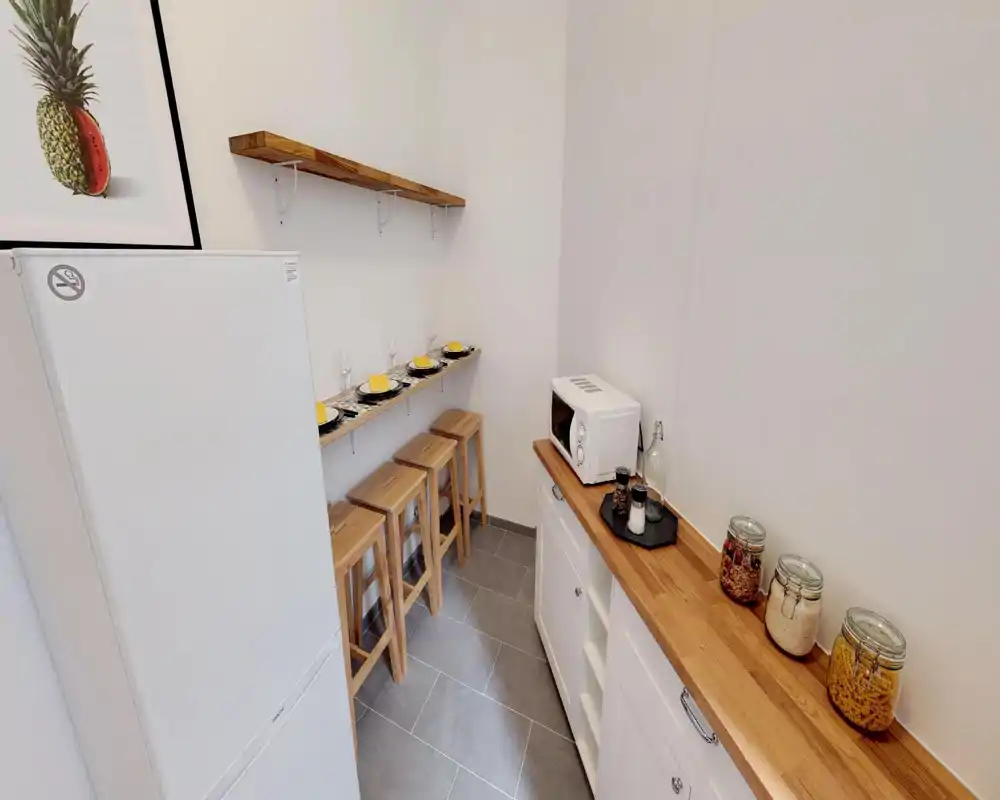 Angolo cottura compatto e moderno con piano in legno, sgabelli e frigorifero. Elettrodomestici e barattoli di vetro creano uno spazio ordinato e funzionale per preparazioni veloci.