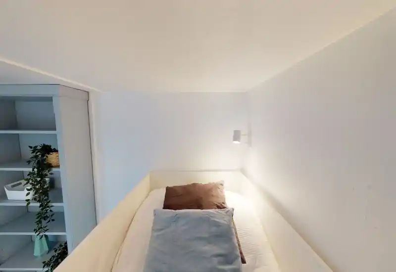 Camera da letto compatta e luminosa con letto singolo incorniciato da pareti bianche e una lampada a muro; tessili essenziali e una scaffalatura vicina offrono comfort e verdura.