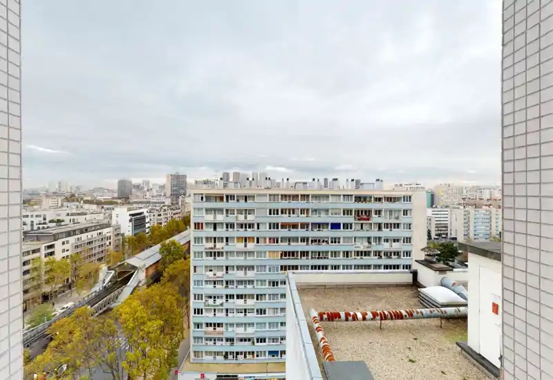 Ampia vista dal tetto/terrazza su un isolato cittadino con facciate di condomini, una strada alberata e una stazione sopraelevata; buona luce diurna e panorama urbano nitido.