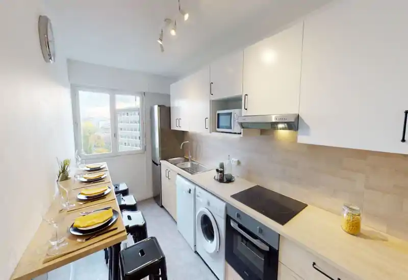 Bright, modern galley kitchen with white cabinets, wood-effect countertops and integrated appliances including oven, hob, fridge and washing machine. A narrow breakfast bar with stools is set for dining by the window.