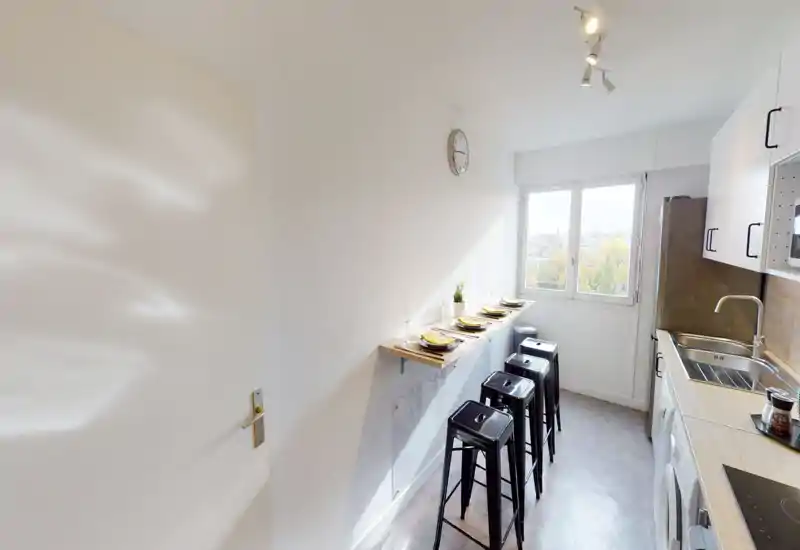 Bright, modern kitchenette with a narrow breakfast bar, four metal stools, built-in sink and appliances, and a window offering outside views.
