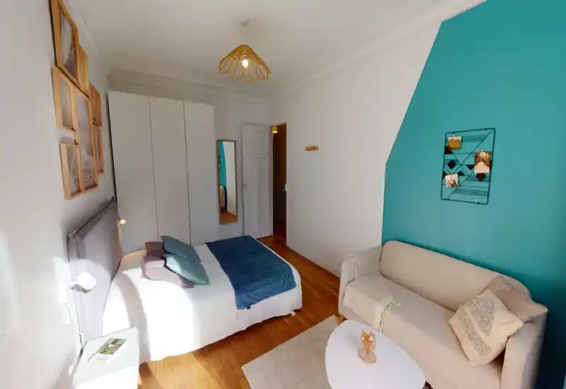 Bright, stylish bedroom with a double bed, bedside table and a comfy sofa against a teal accent wall. Soft linens, decorative cushions and warm wood floors create a welcoming atmosphere ideal for short-term rentals.