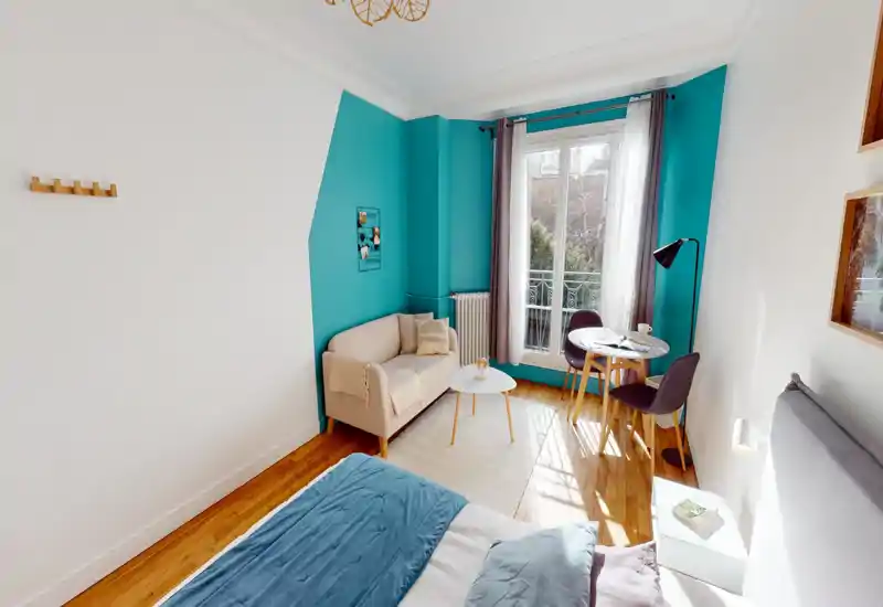 Bright, styled bedroom with a visible double bed in the foreground, a small beige sofa, round coffee table and a dining nook by a large window offering pleasant natural light.