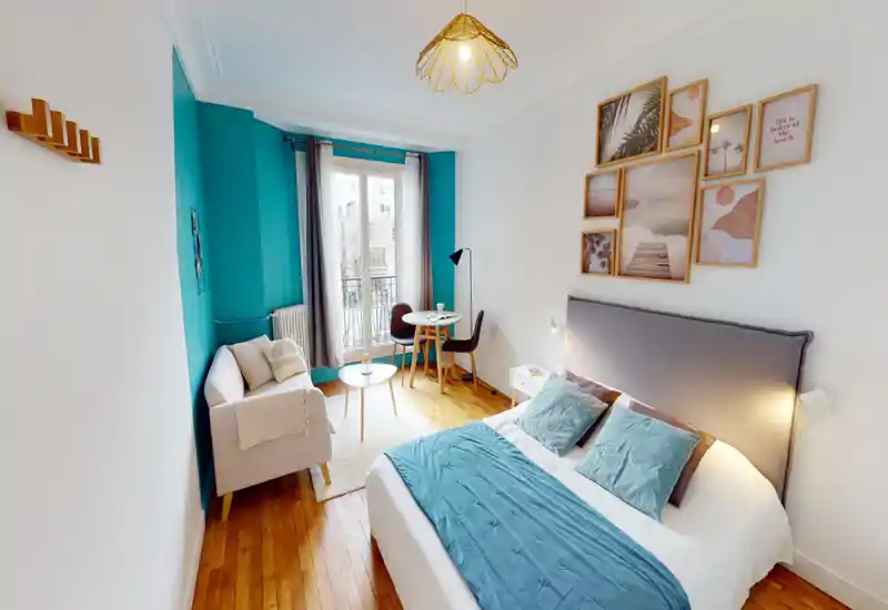 Bright, stylish bedroom with a comfortable double bed, soft teal accents and coordinated wall art. The room features wooden floors, a small seating area and a table by a large window that provides natural light.