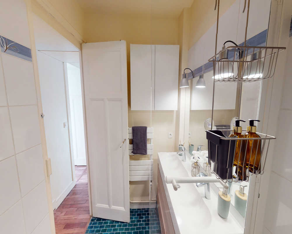 Clean, well-lit bathroom with double sink vanity, wall-mounted cabinets, mirror and organized soap dispensers; tiled floor and towel radiator visible.