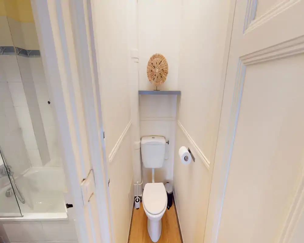 Compact, well-maintained toilet room with a standard WC, toilet brush, and wall shelf; bright, neutral decor and wood-effect flooring.