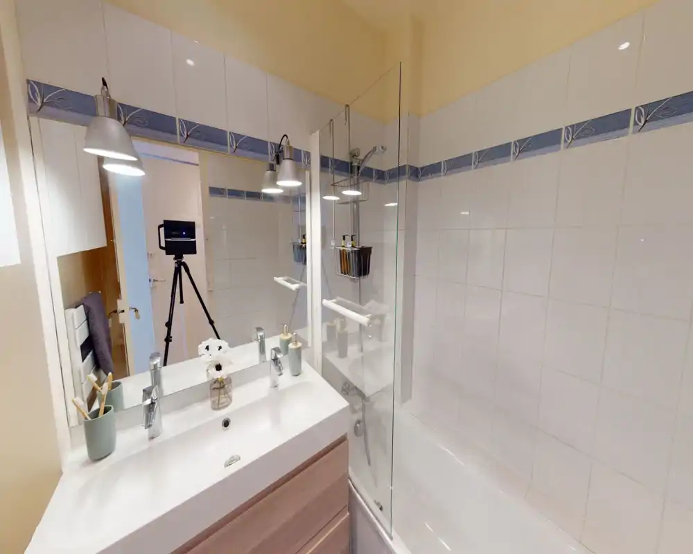 Bright, well-lit bathroom with a sink and bathtub/shower combination, white tiles with decorative blue border, modern chrome fixtures and a large mirror.