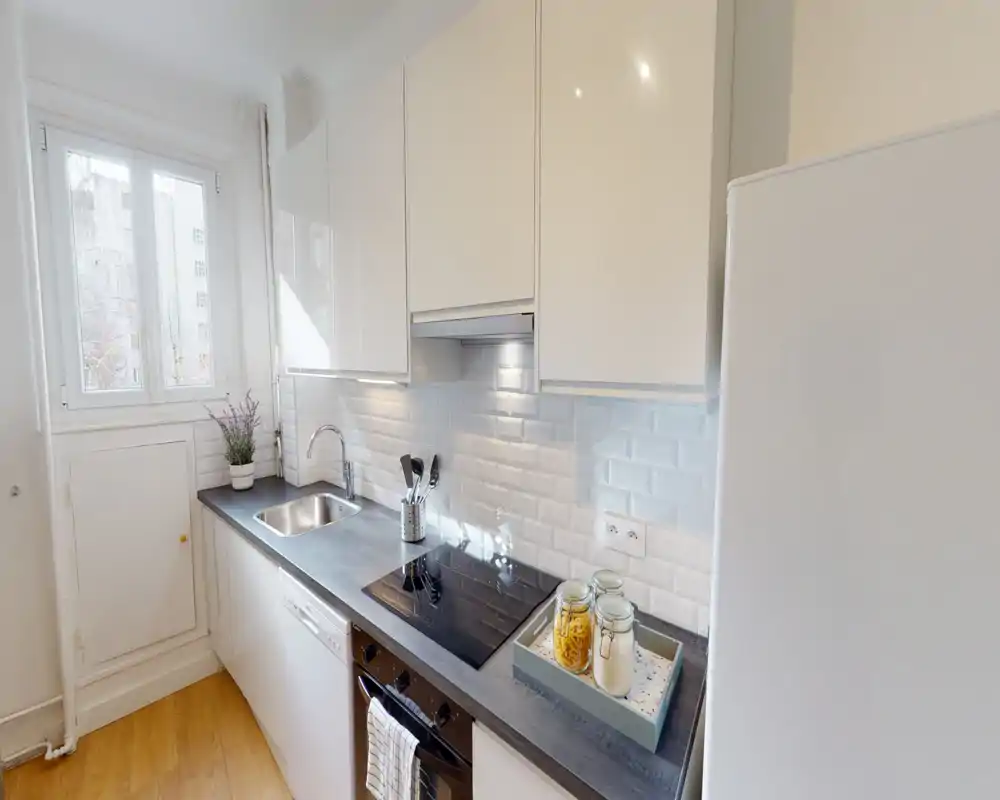 Bright modern kitchen with white high-gloss cabinets, a dark countertop, integrated hob and oven, and a stainless sink. Natural light from a window and small decorative details (plant, jars) create a fresh, tidy space.
