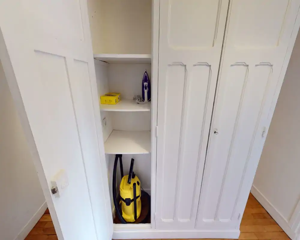 Small built-in storage/closet near an entrance with shelves holding a vacuum cleaner and iron; white cabinetry and wooden floor.