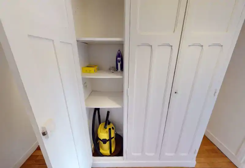 Small built-in storage/closet near an entrance with shelves holding a vacuum cleaner and iron; white cabinetry and wooden floor.
