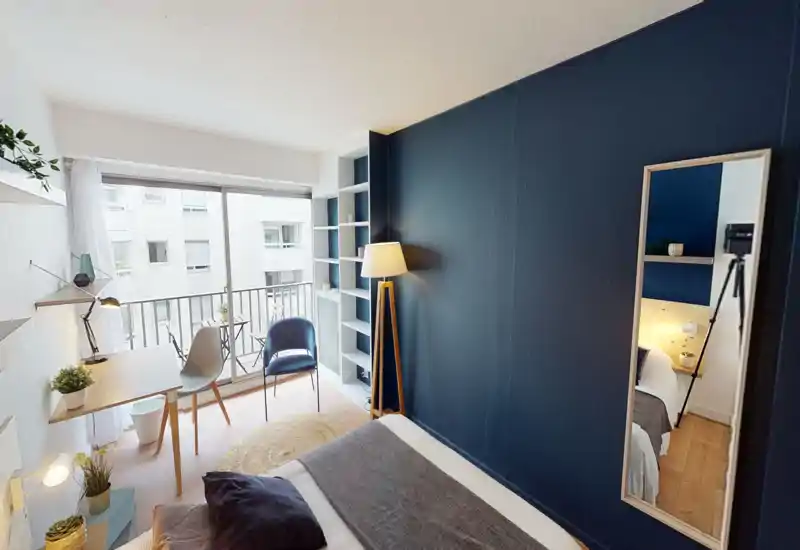 Bright, modern bedroom with a neatly made bed in the foreground, a desk by the window and a cozy reading corner with a floor lamp and chair.