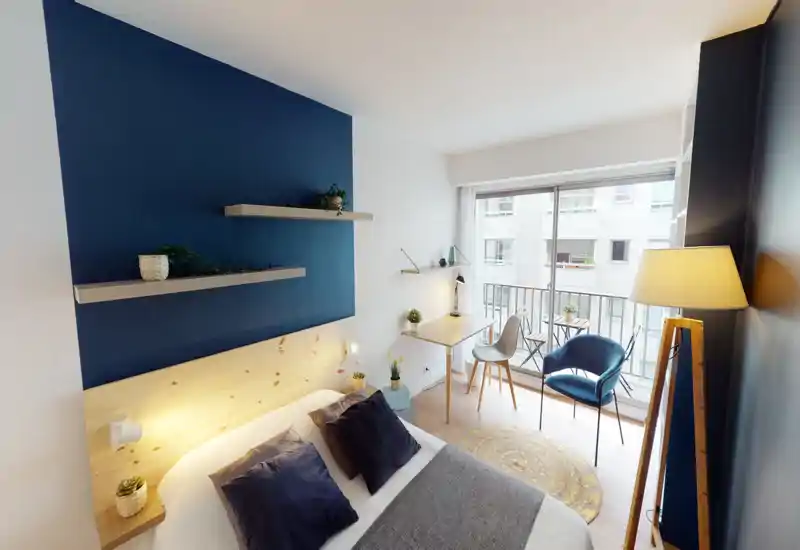 Bright, stylish bedroom with a large double bed, layered cushions, wooden headboard and a desk by floor-to-ceiling windows opening onto a small balcony. Contemporary navy accent wall, decorative shelving and warm lighting create a cozy, well-styled space ideal for short stays or city rentals.