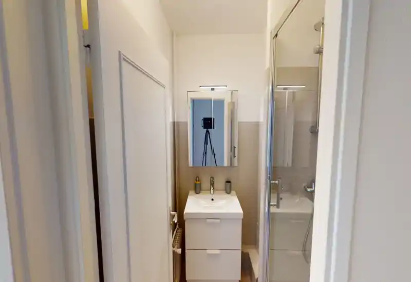Compact, modern bathroom with a white vanity, sink and mirrored cabinet; glass shower door visible on the right and neutral beige wall tiles.