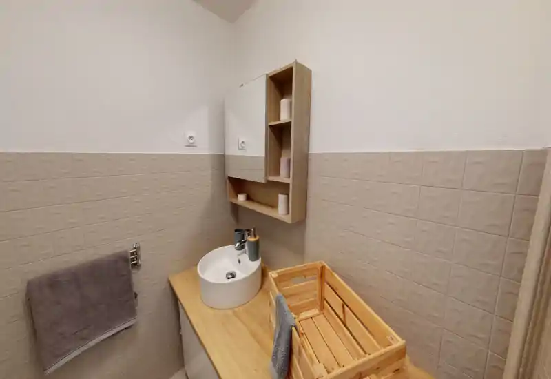 Compact, modern bathroom with a round countertop sink, wood vanity and wall-mounted mirrored cabinet. Neutral textured tiles and tidy accessories create a clean, functional space.
