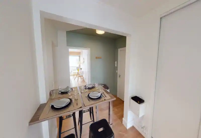 Cozy, staged dining area with a narrow wooden table set for four and high stools; bright connection to an adjacent living space provides natural light.