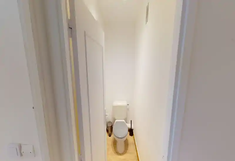 Compact private toilet with white walls, wall-mounted cistern and a wooden-effect floor. Clean, simple and well lit — ideal as a guest WC.