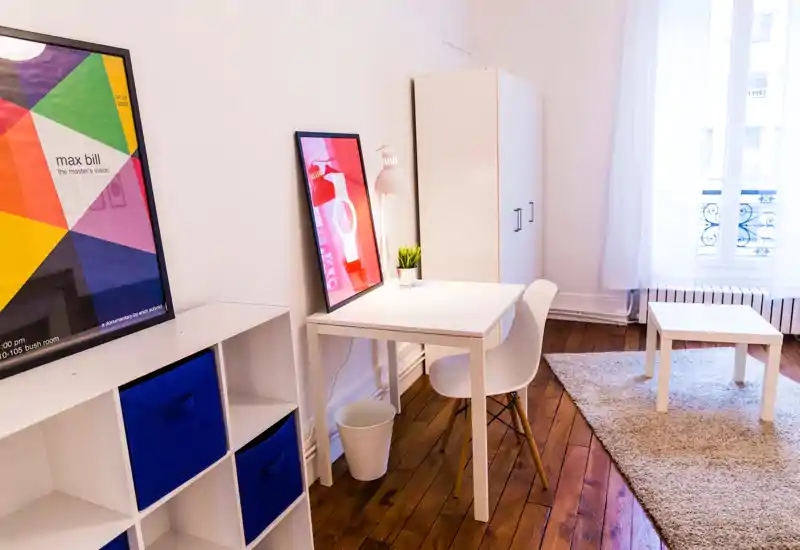 Espace de vie lumineux et soigné avec petite table, chaise type fauteuil, étagère à cases avec bacs en tissu et cadres décoratifs — parquet chaleureux et tapis moelleux pour une ambiance accueillante.