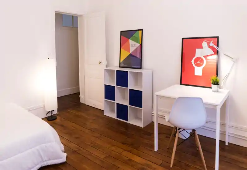 Chambre lumineuse et minimaliste avec un coin de lit visible, parquet, petit bureau et étagères à cubes — propre, moderne et confortable pour une personne.