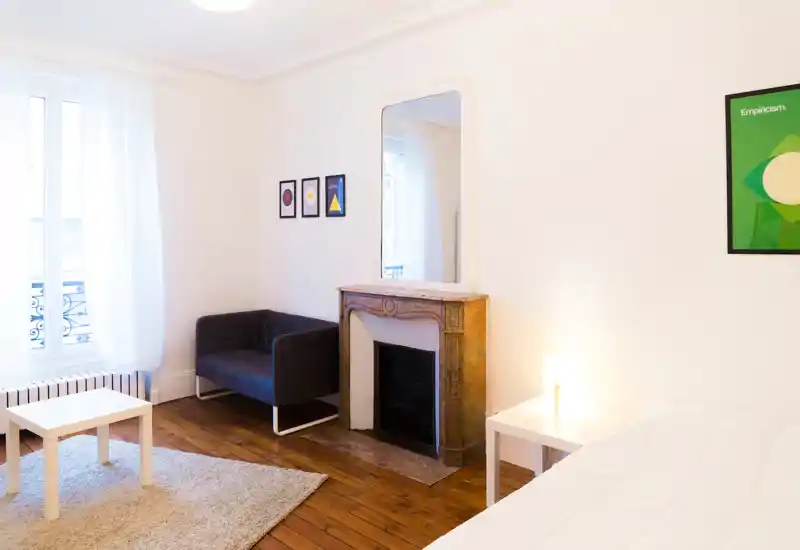 Coin de chambre lumineux et minimaliste avec une petite partie du lit visible, un tapis douillet, un petit canapé sombre et une cheminée décorative surmontée d’un grand miroir — idéal pour des annonces au style épuré et aéré.