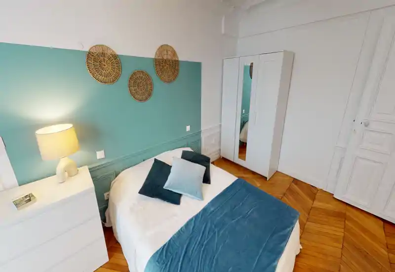 Bright, stylish bedroom with a double bed dressed in white linens and teal accents, decorative wall pieces, a bedside lamp, chest of drawers and a wardrobe — warm wooden herringbone floor.