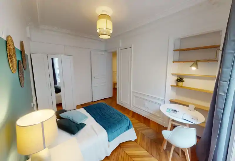 Bright, stylish bedroom with a comfortable double bed dressed in teal and white linens, parquet floors and a small workspace by the window. The room features built-in shelving, a wardrobe with mirrored door and warm accent lighting — ideal for a short-term rental.