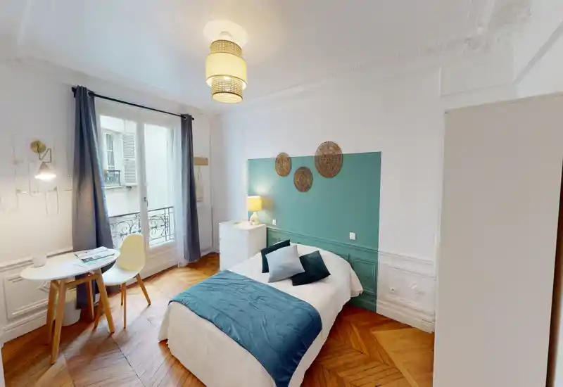 Bright, stylish bedroom with decorative molding, teal accent wall and a comfortable double bed dressed in coordinated linens. Natural light from a French window, wooden herringbone floor and a small work table make this room ideal for both rest and remote work.